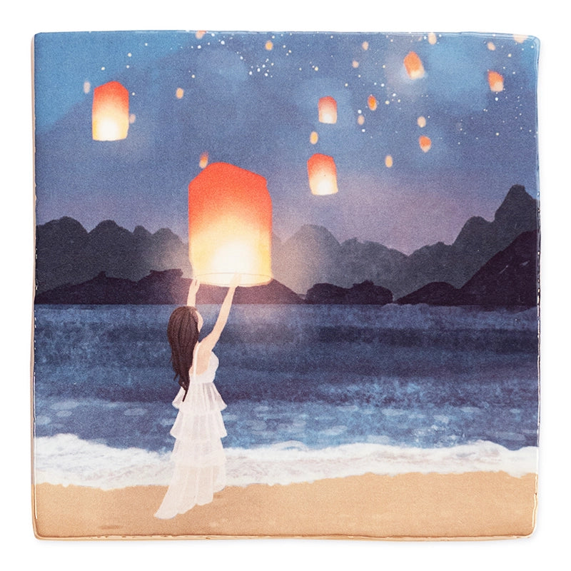 StoryTile Making memories & wishes. Op dit tegeltje staat een vrouw in witte jurk die een wetsballon oplaat. Ze staat op het strand aan de rand van de zee. op de achtergrond zie je bergen. Overal in de lucht vliegen wetsballonnen. 