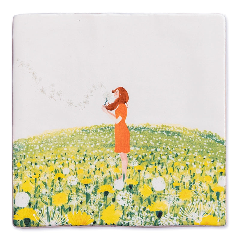StoryTile Ik heb een wens. Prachtige decoratietegel met daarop een jonge vrouw in oranje jurk, met rode haren in een veld vol paardebloemen. Ze heeft een bloem vast en blaast de pluisjes weg in de wind. 