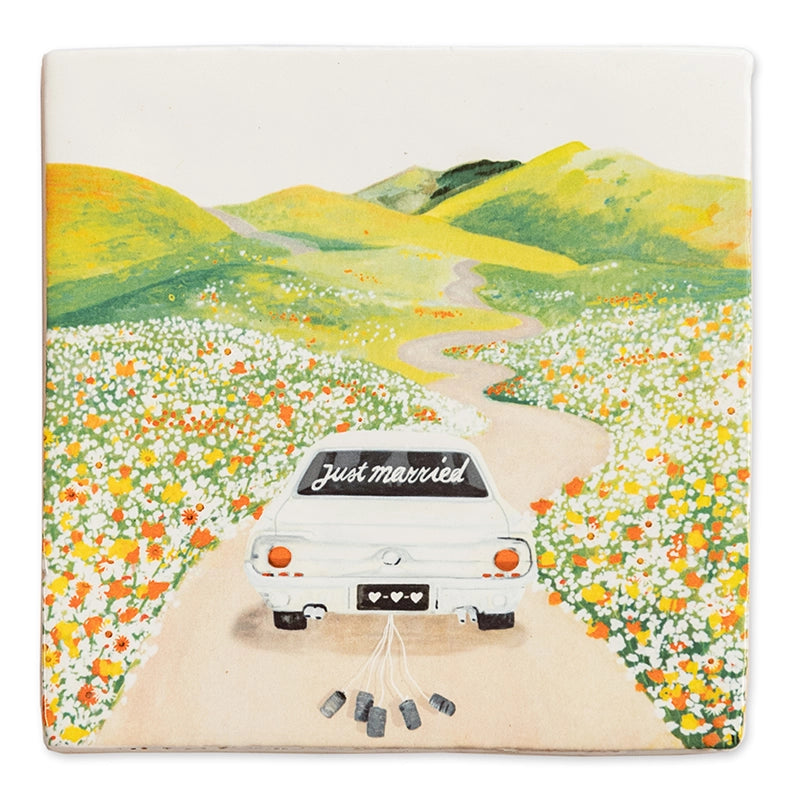 StoryTile - Just married. Net getrouwd! Deze StoryTile toont een witte auto met op de achterruit de woorden 'Just married'. Achter de auto hangen lege blikjes. De auto rijdt door een heuvellandschap met bloemen in het veld.  